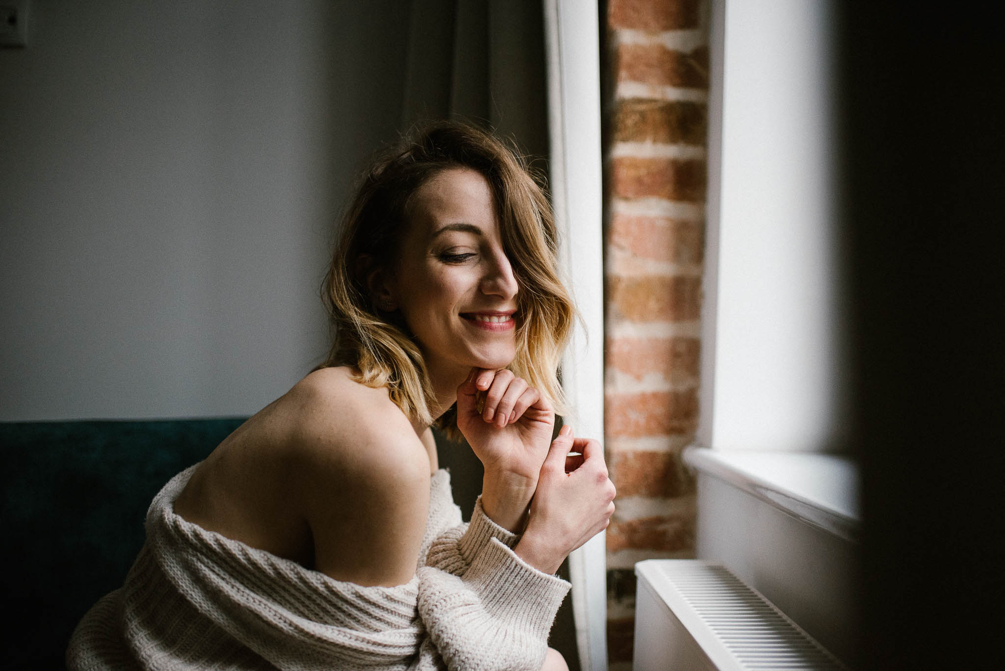Kobieta w&nbsp;swetrze pokazuje ramię i&nbsp;siedzi przy oknie - sesja kobieca w&nbsp;apartamencie w&nbsp;Poznaniu