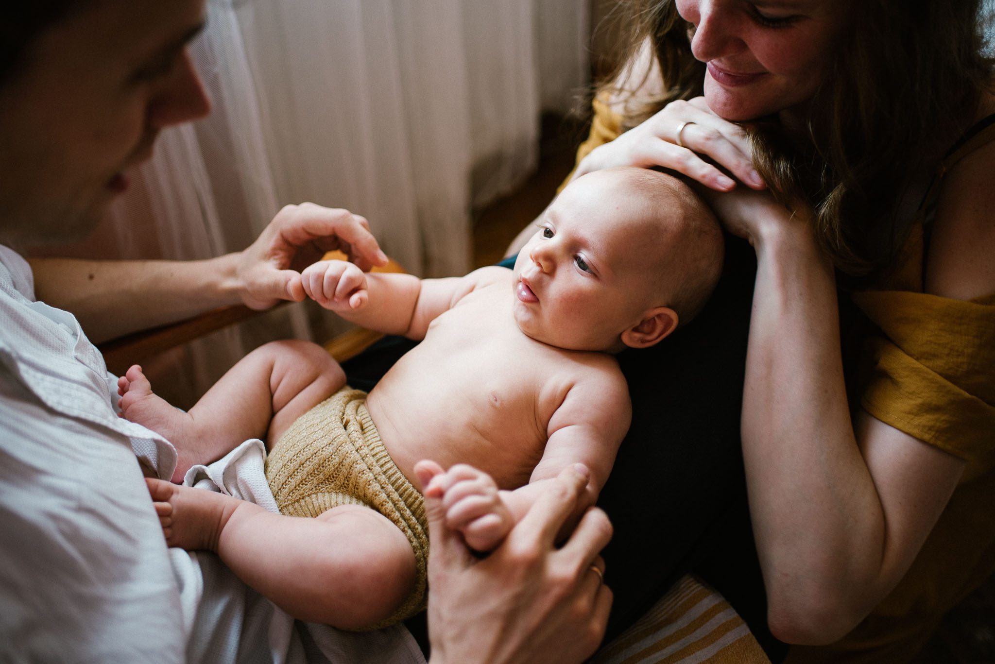 Tata trzyma niemowlę na&nbsp;kolanach, mama siedzi obok nich -Sesja niemowlęca lifestyle w&nbsp;domu Poznań