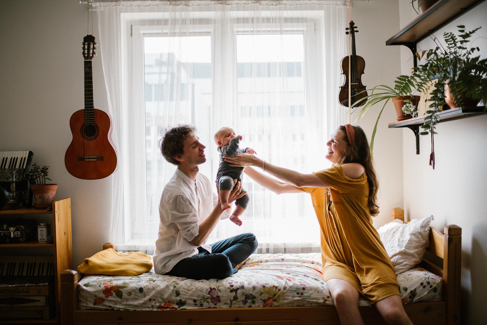 tata przekazuje niemowlę mamie na&nbsp;łóżku - Sesja niemowlęca lifestyle w&nbsp;domu Poznań